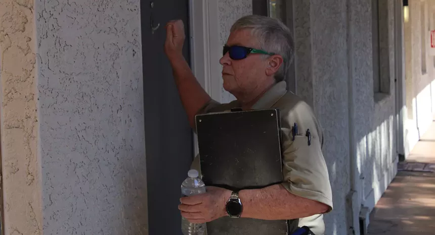 a woman with short gray hair and sunglasses and a tan polo knocks on a door while holding a clipboard