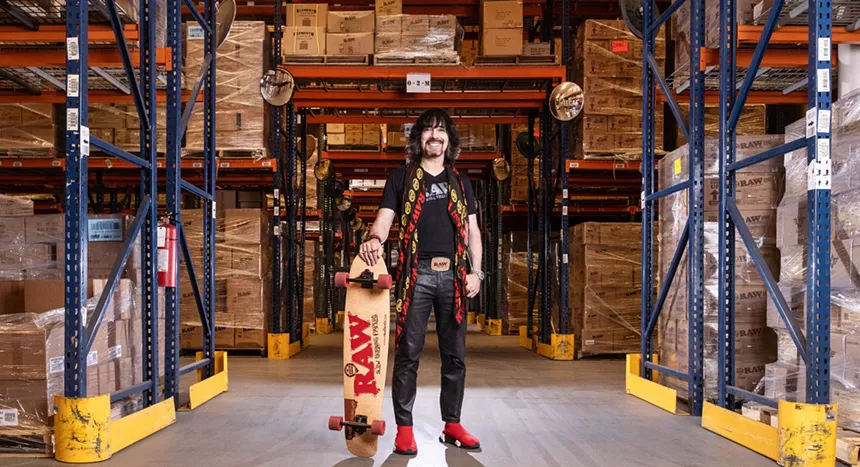 a man holds a skateboard that says "RAW" inside a warehouse stacked with boxes on shelves