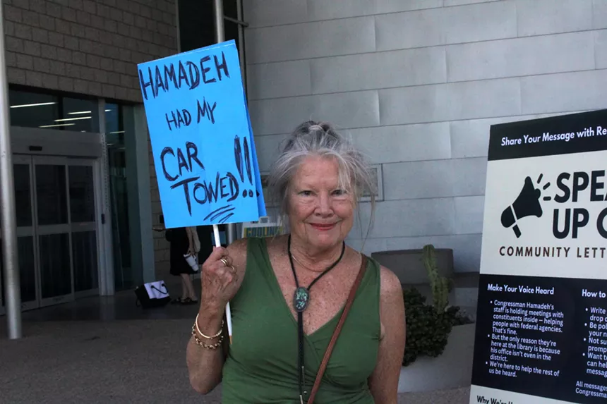 a woman holds a blue sign that says "hamadeh had my car towed"