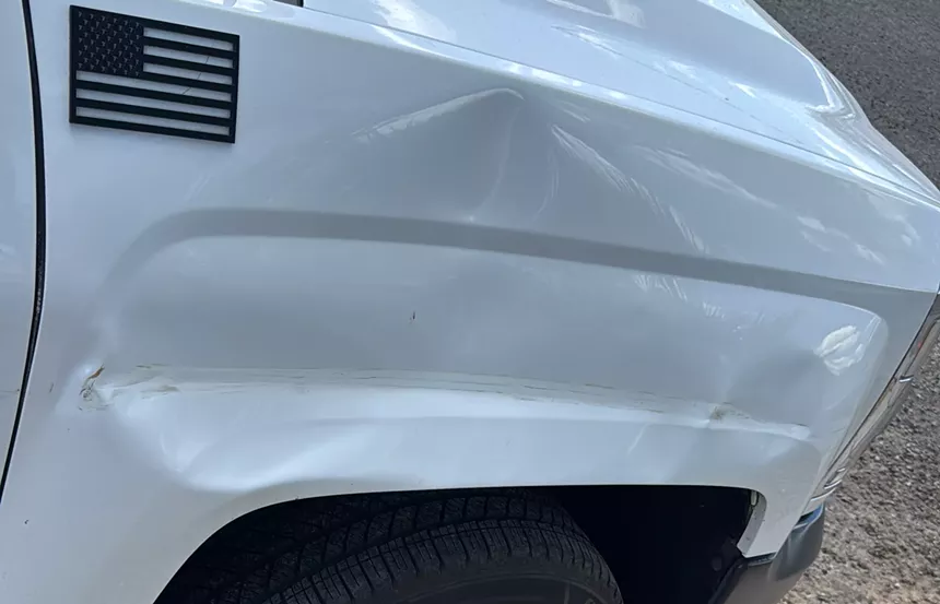 the front passenger side of a white truck with a big dent. an american flag decal is visible