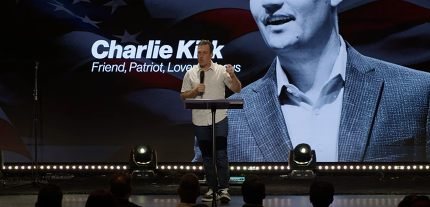 a man stands on a stand in front of an image of charlie kirk