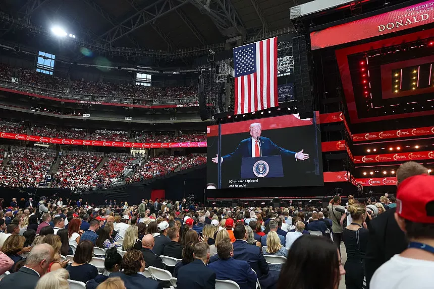 attendees at a large stadim watch donald trump on a big screen