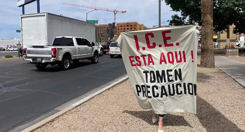 Image: A woman holds up a large banner that reads: “ICE is here! Take precautions,” in Spanish at the Phoenix Immigration Court in Phoenix